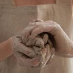 Unrecognizable person wearing gray apron shaping and molding clay in dirty hands while making handicraft crockery in workshop during work